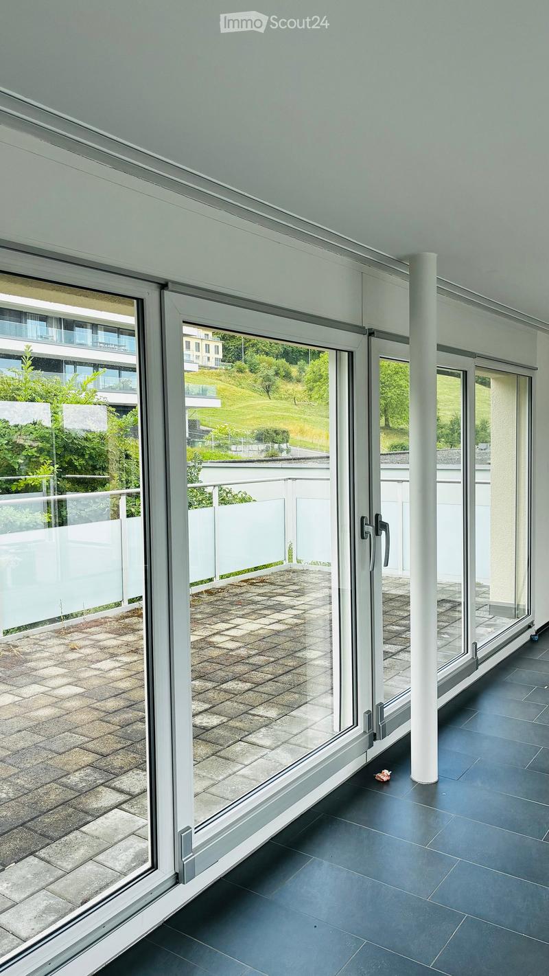 Modern balcony with large glass sliding doors, dark tiled floor, and a view of a green hillside and neighboring buildings.