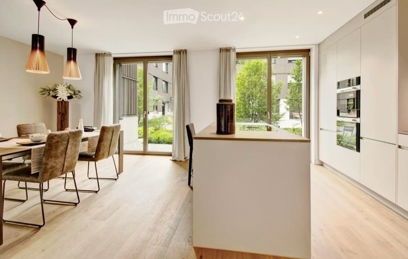 Open-plan kitchen and dining area with light wood flooring, white cabinetry, modern pendant lights, and a dining set with grey chairs.
