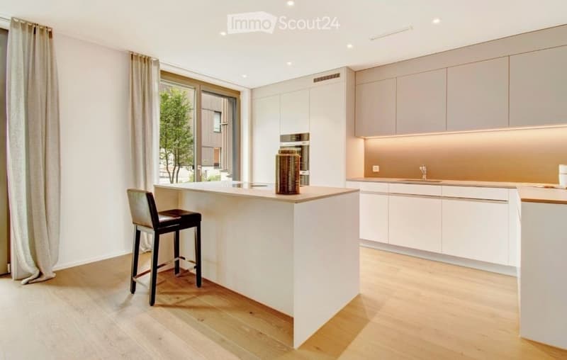 Modern kitchen with white cabinets, a central island, a bar stool, and a decorative vase on the island. The space features wooden flooring and large windows with curtains.