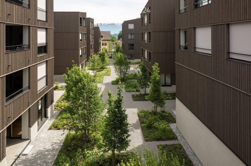 Courtyard view of a modern residential complex with brown wooden facades, balconies, and landscaped greenery including trees and shrubs.