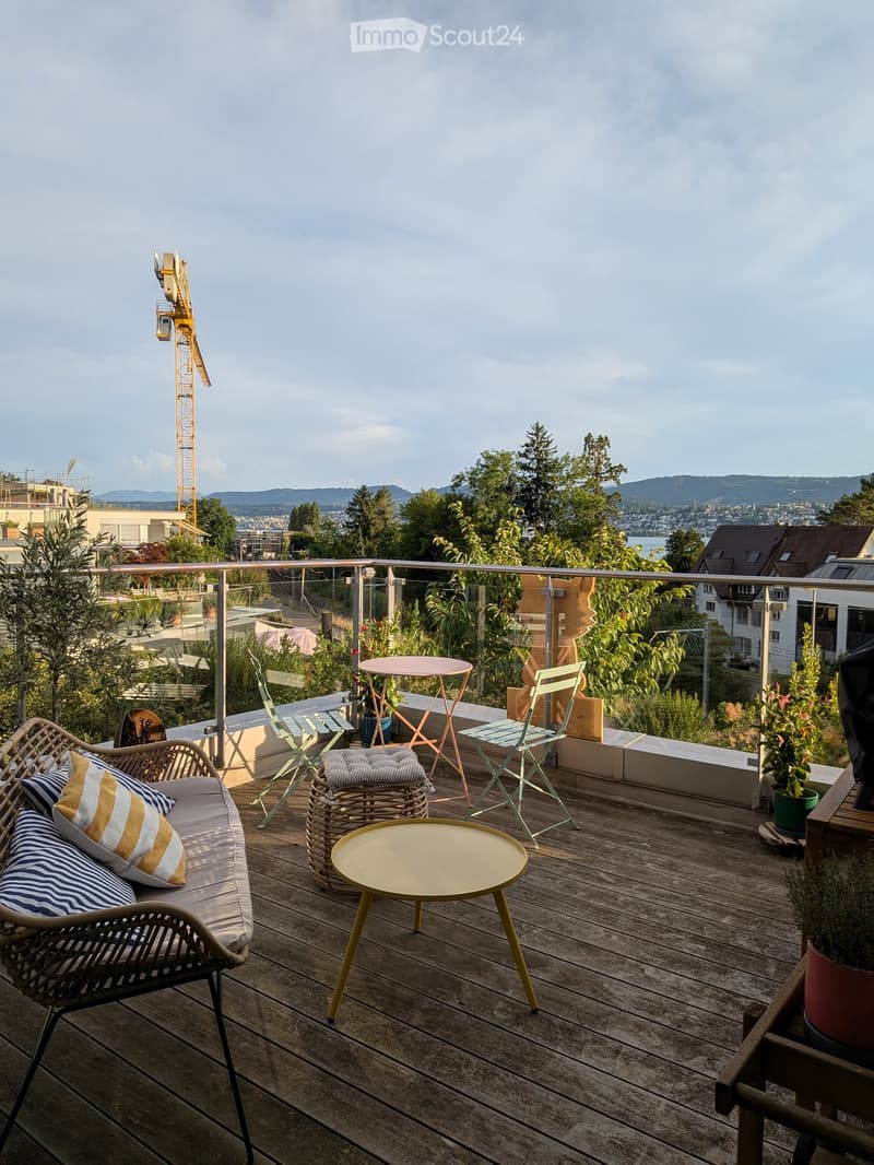 Wooden deck with a sofa, small tables, chairs, and potted plants, overlooking a cityscape with trees and a crane in the distance.