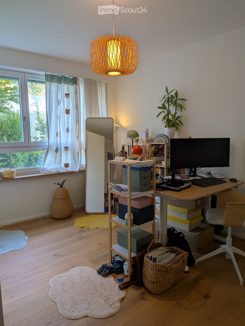 A well-lit room with wooden flooring, a large window with curtains, a desk with a computer setup, a shelf with storage boxes, a mirror, and decorative items including a plant and a wicker basket.