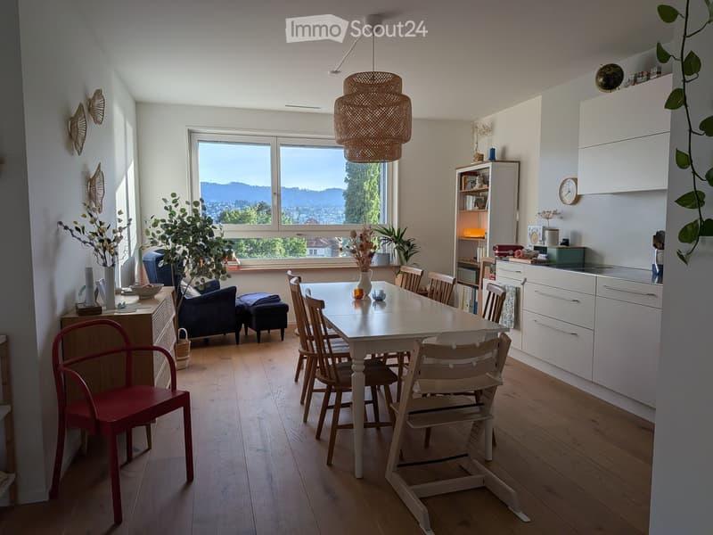 Bright, open-plan living and dining area with wooden flooring, large windows offering a scenic view, a dining table with chairs, a sofa, a stool, a kitchen area with cabinets, a bookshelf, and decorative plants.