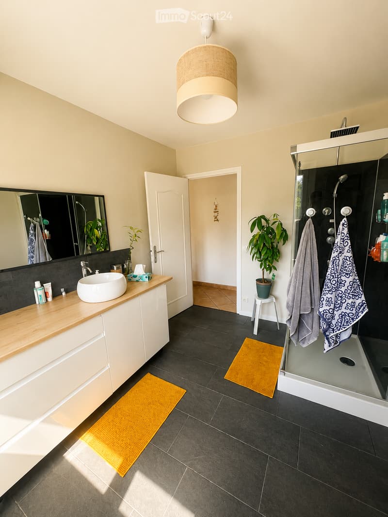 Modern bathroom with a large sink, mirror, and a glass-enclosed shower. Features include a wooden countertop, dark tiled floor, and decorative yellow rugs.