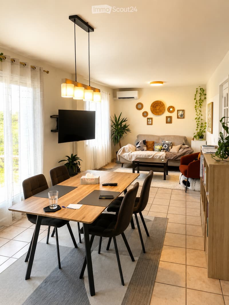 A modern living room with a sofa, coffee table, dining table with chairs, a TV screen mounted on the wall, and decorative plants. The space features a mix of wooden and tiled flooring, with large windows covered by sheer curtains.