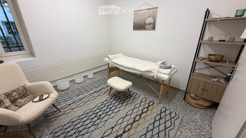 A well-lit room with a massage table, a comfortable sofa, a small stool, shelves with decorative items, and a painting on the wall.