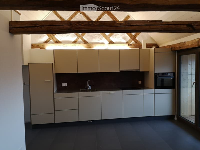 Modern kitchen with light-colored cabinets, stainless steel sink, and built-in oven. Exposed wooden beams on the ceiling and dark tiled floor.