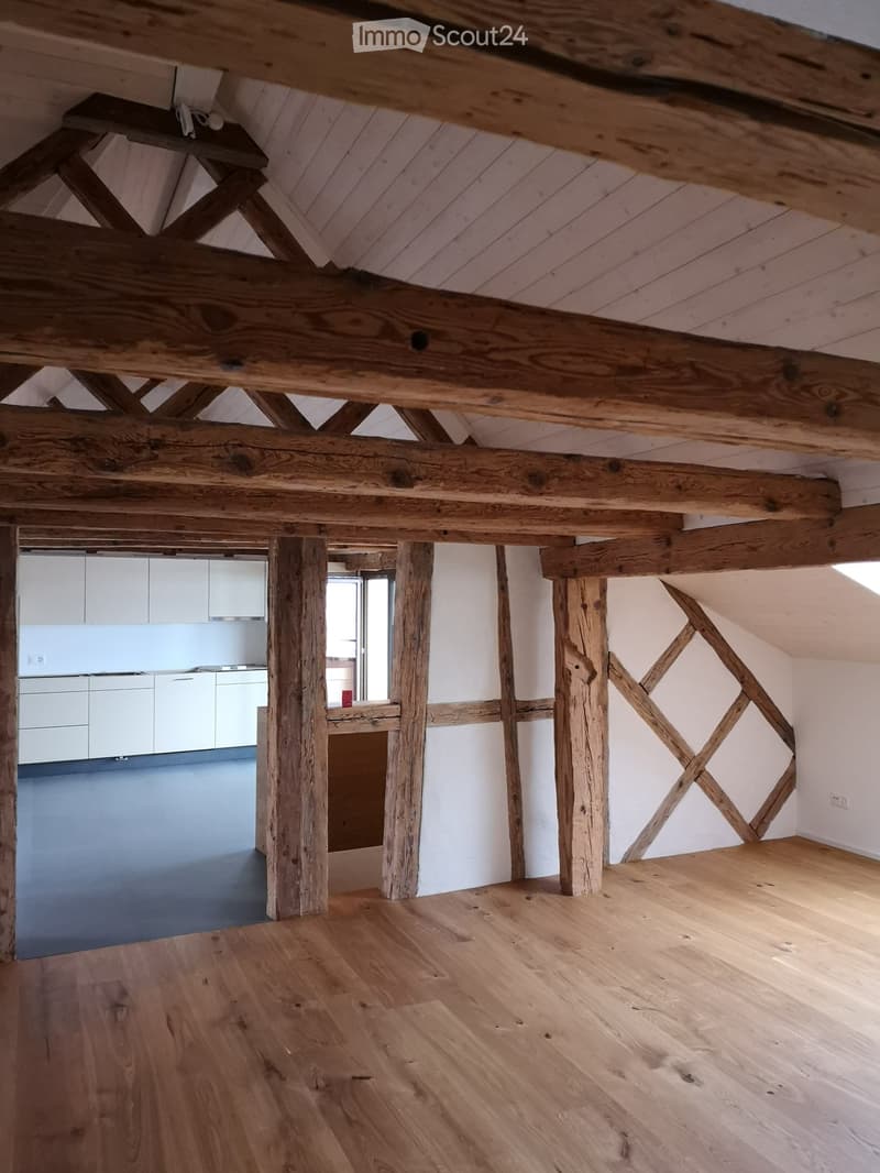 Loft-style room with exposed wooden beams and pillars, wooden floor, white walls, and a modern kitchen area with white cabinets and a countertop.
