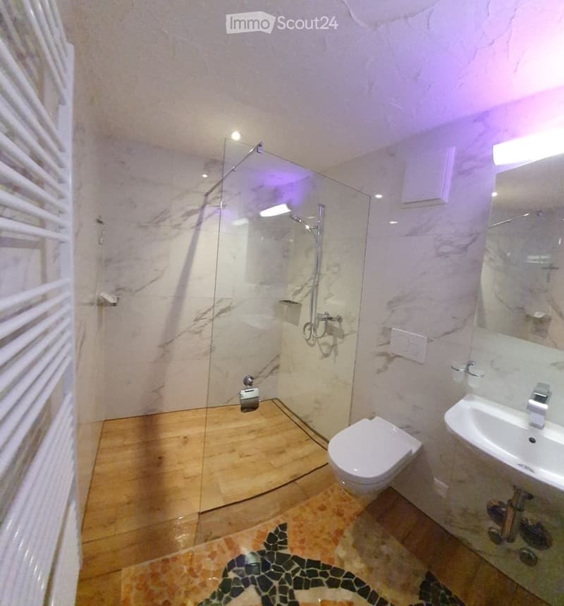 Modern bathroom with marble walls, wooden floor, glass-enclosed shower, toilet, and sink.