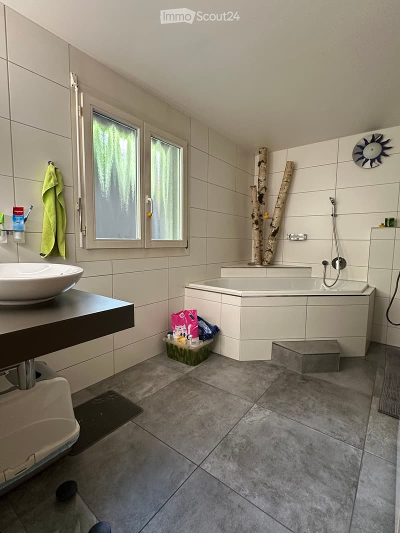 Modern bathroom with a large bathtub, separate shower, sink, and a window. Includes a towel, toothbrush, toothpaste, and a cat litter box with cat food.