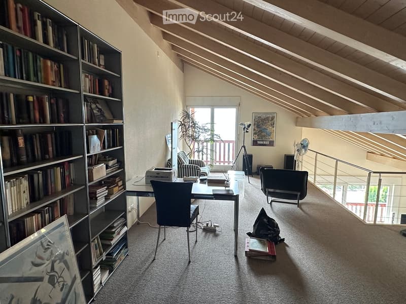 A well-lit attic room with a slanted wooden ceiling, featuring a large bookshelf filled with books, a desk with a chair, a printer, and a fan. There is a tripod near a window with a view, and a small seating area with a chair and a plant.