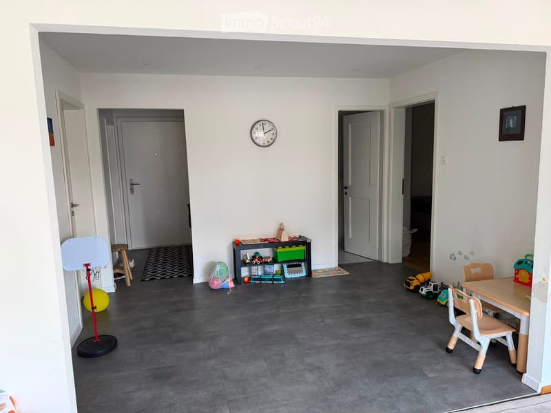 A well-lit, open-plan room with grey tiled flooring, white walls, and several doors leading to other areas. The room contains a small table with chairs, a shelf with toys and storage boxes, a basketball hoop, a ball, and a clock on the wall.