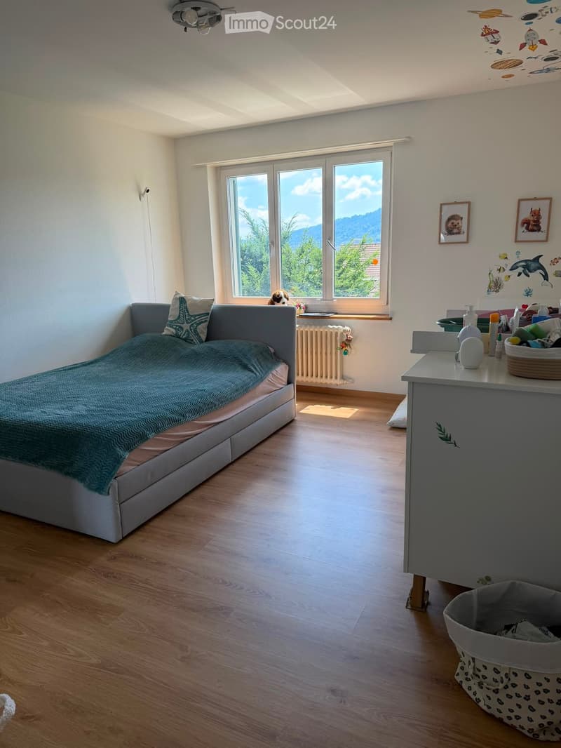 Bright bedroom with wooden flooring, large windows offering a scenic view, a grey sofa with a teal blanket, a small table with various items, and a framed picture on the wall.