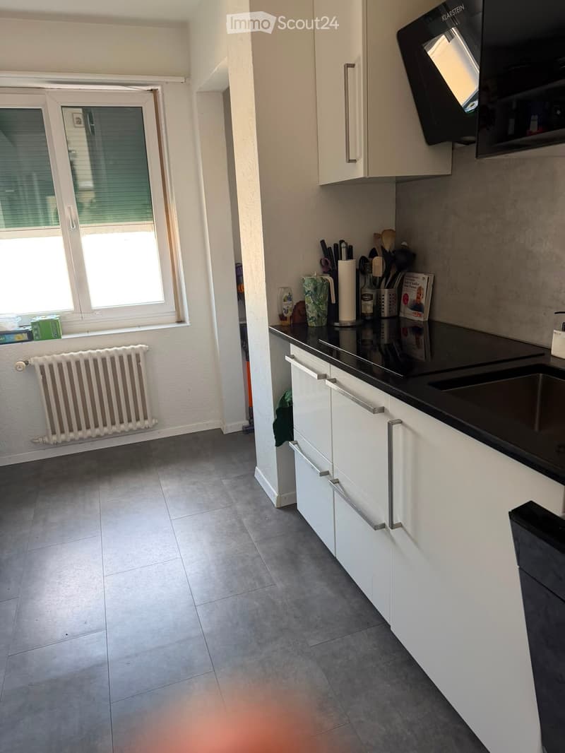 Modern kitchen with white cabinets, black countertop, stainless steel sink, microwave, and a window with blinds. The floor is tiled, and a radiator is visible on the wall.