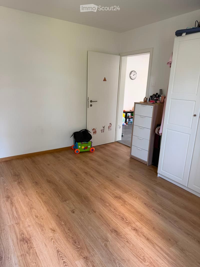 A small, well-lit room with wooden flooring, a white door, a drawer unit, a toy car, and a backpack. The room has a clock on the wall and a partially visible adjacent room with more furniture.