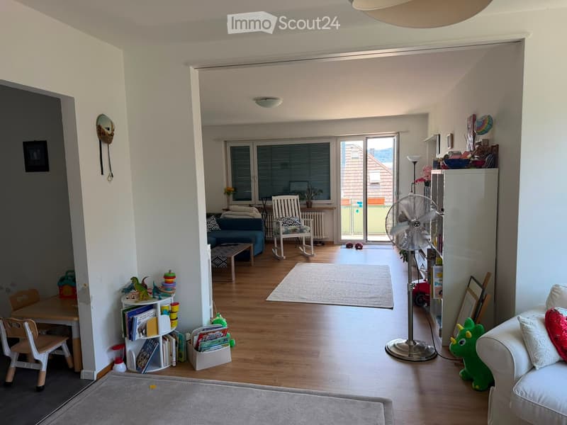 A well-lit living room with wooden flooring, a sofa, a coffee table, a fan, a bookshelf, and various toys and decorations.