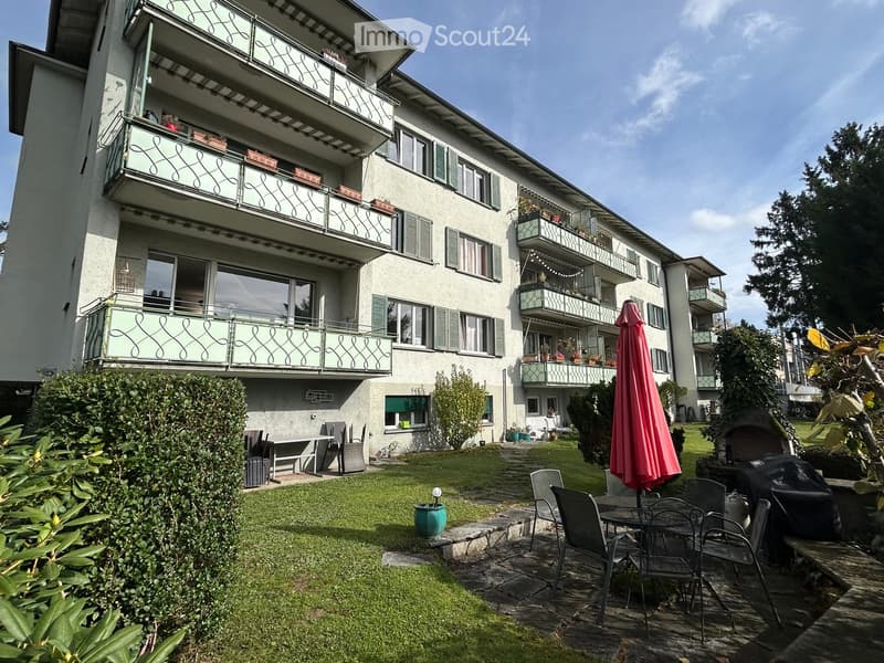 Garden area with a patio, outdoor furniture, and a closed umbrella, adjacent to a multi-story building with balconies.
