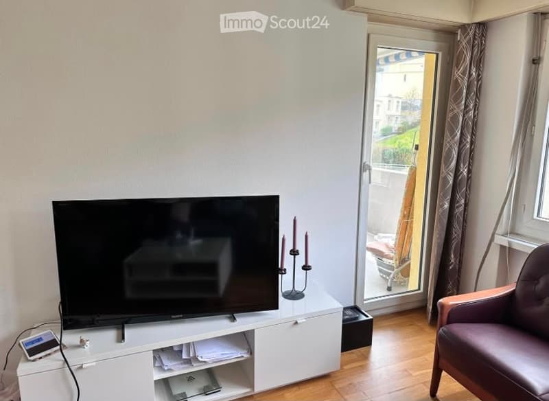 A modern living room with a flat-screen TV on a white cabinet, a purple armchair, and a candle holder on the table. The room has wooden flooring and large windows with curtains.