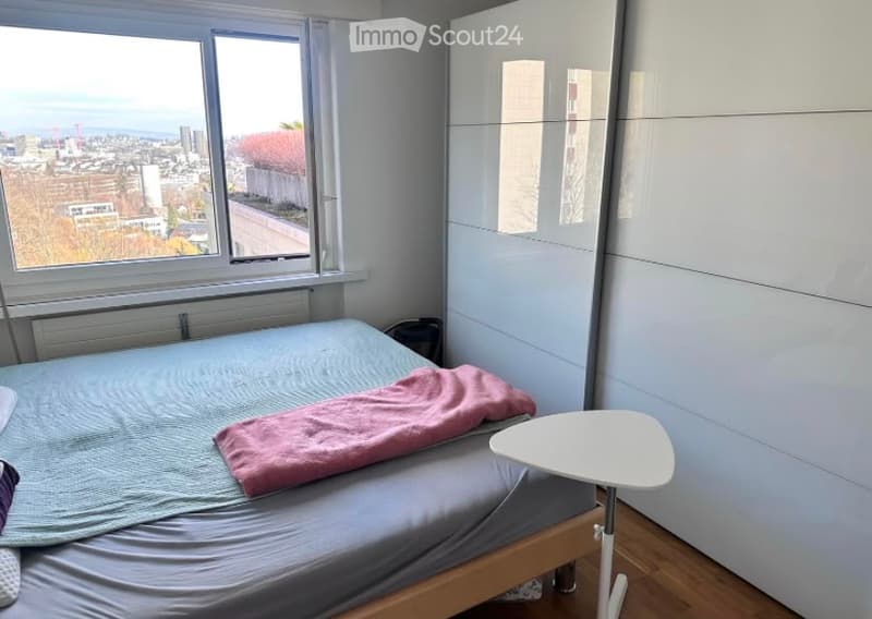 Modern bedroom with a double bed, nightstand, and large wardrobe. The room features large windows offering a city view.