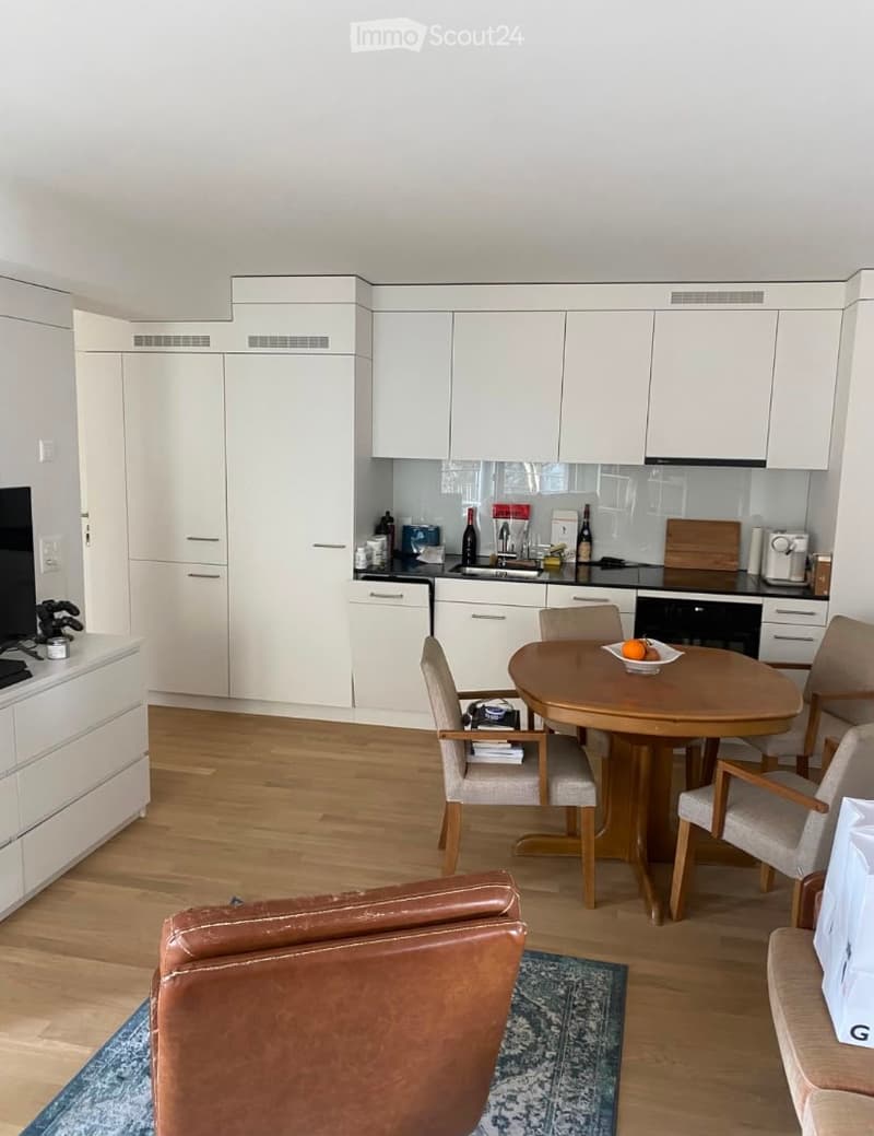 A modern, open-plan living and dining area with white cabinetry, a wooden dining table with chairs, a leather sofa, and a kitchen area featuring a dishwasher, refrigerator, and microwave.