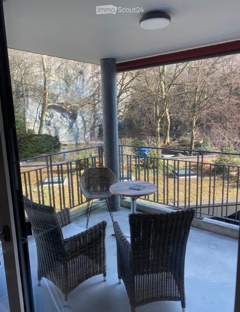 Balcony with a round table and two wicker chairs, overlooking a park with trees and a body of water.