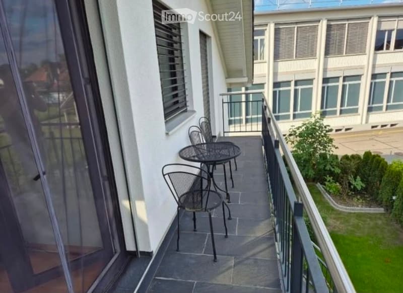 Balcony with a small table and chairs, overlooking a landscaped area with greenery and a building in the background.