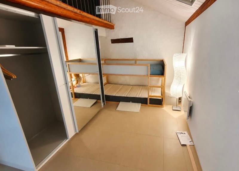 A compact room with a wooden bunk bed, a wardrobe, and a modern floor lamp. The room features light-colored tiled flooring and white walls.