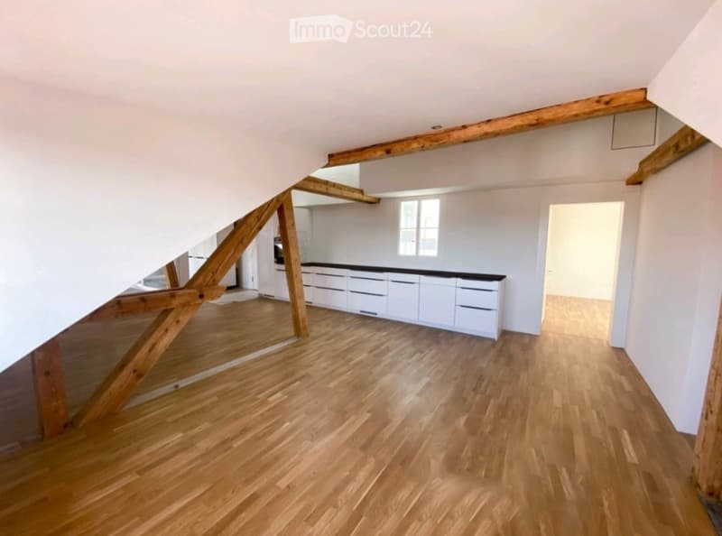 An unfurnished attic room with wooden beams, wooden flooring, and white walls. A doorway leads to a kitchen area with white cabinets and a black countertop.