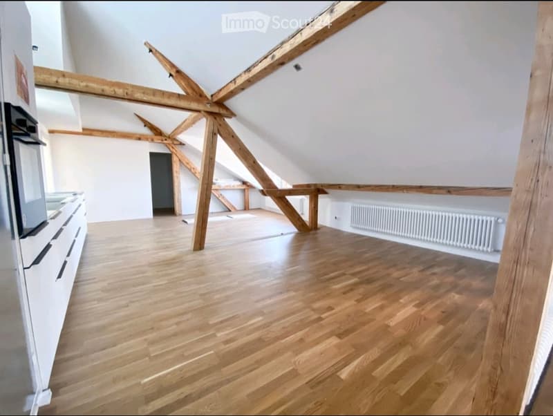 Empty attic space with exposed wooden beams, wooden flooring, white walls, and a radiator. No furniture or equipment present.