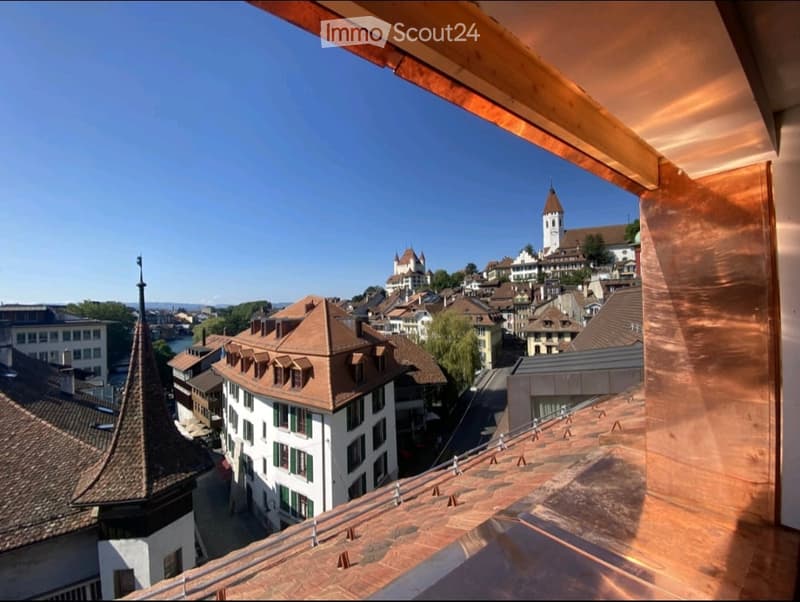 View from a balcony or terrace overlooking a historic town with traditional buildings, a church tower, and a clear blue sky.