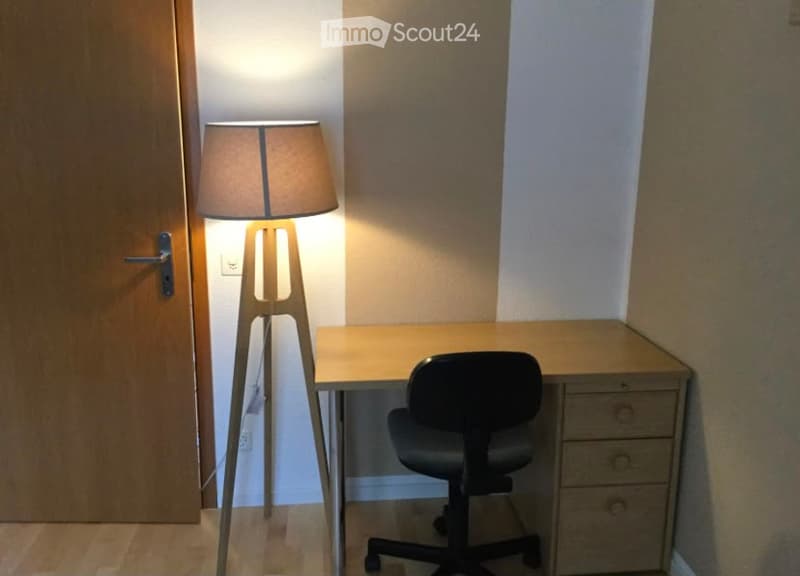 A small, modern workspace with a wooden desk, black office chair, and a tall floor lamp with a beige shade. The room has light-colored walls and a wooden door.