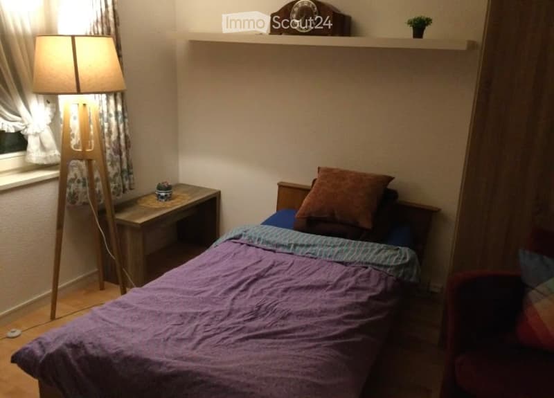 Cozy bedroom with a single bed, nightstand, chair, table lamp, shelf with clock and plant, and a small wooden desk with a lamp.