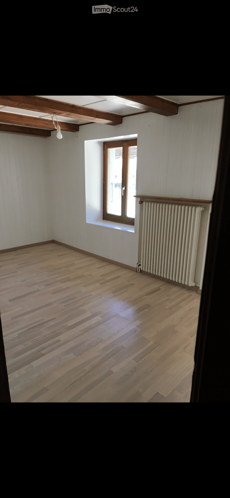 Empty room with wooden flooring, white walls, exposed wooden beams on the ceiling, a radiator, and a window with a wooden frame.
