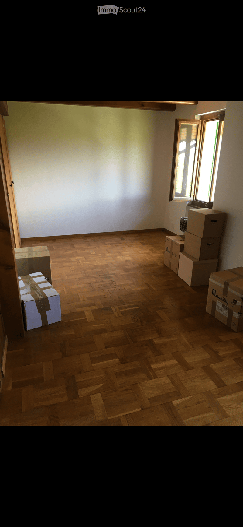 Empty room with wooden herringbone flooring, white walls, and a window with wooden frame. Several cardboard boxes are stacked in the corners.