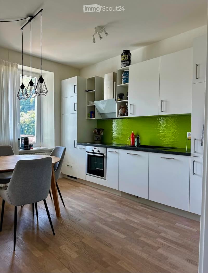 Modern kitchen with white cabinets, a green backsplash, and a wooden dining table with chairs. Features an oven, countertop, and pendant lighting.