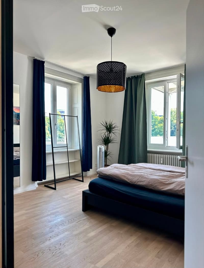 Modern bedroom with wooden flooring, large windows, dark curtains, a bed with a beige duvet, a hanging lamp, a plant, and a wardrobe.