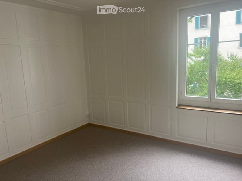 Empty room with white paneled walls, carpeted floor, and a large window with a view of greenery.