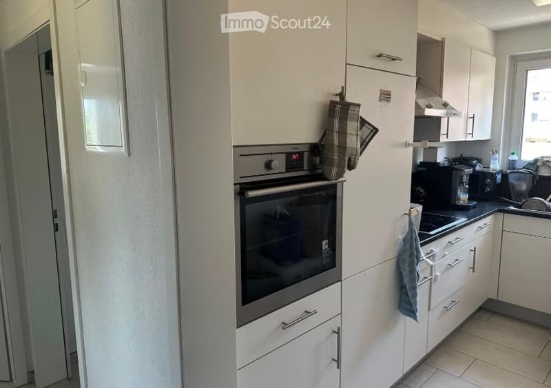 Modern kitchen with white cabinets, stainless steel oven, coffee maker, toaster, and various kitchen appliances. A dish rack is visible, and a plaid oven mitt hangs on the cabinet.