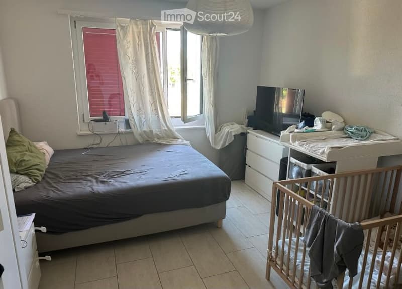 A well-lit bedroom with a bed, pillows, a wooden cradle, a wardrobe, and a television. The room features tiled flooring and a window with white curtains.
