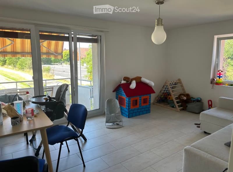 Bright living room with large sliding glass doors leading to a balcony, a sofa, a small table with chairs, a baby chair, a toy house, and various toys including a ladder and stuffed animals.