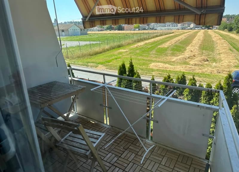 Balcony with wooden table and chairs, overlooking a rural landscape with fields and trees.
