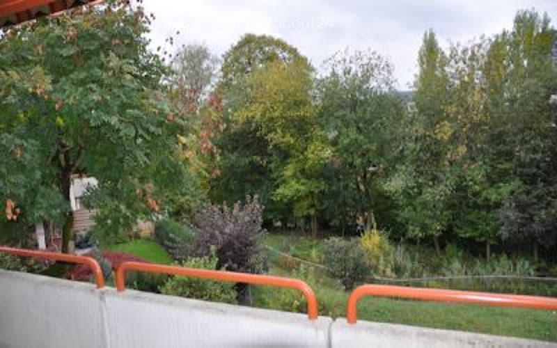 A serene garden area with lush green trees, shrubs, and a well-maintained lawn, enclosed by a concrete wall with orange railings.
