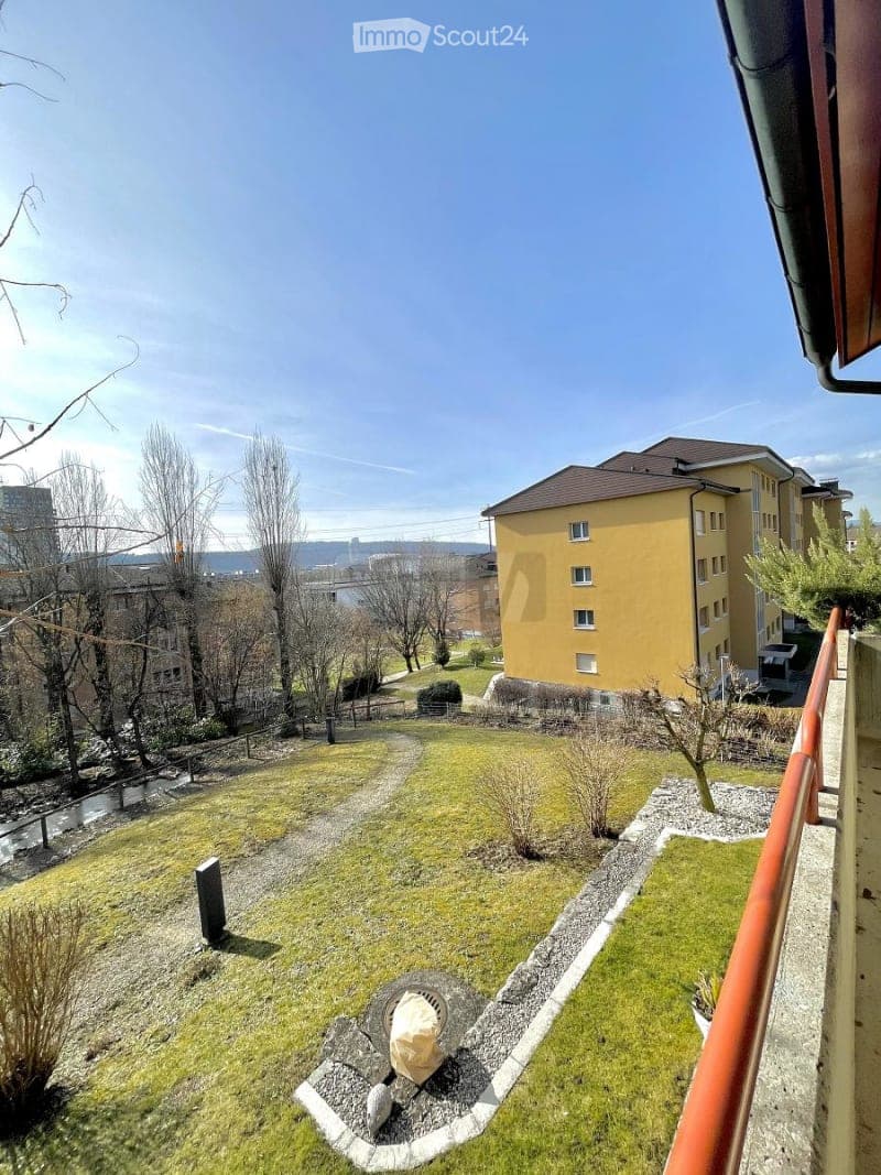 Garden area with grass, gravel pathways, small plants, and a view of a yellow apartment building in the background.