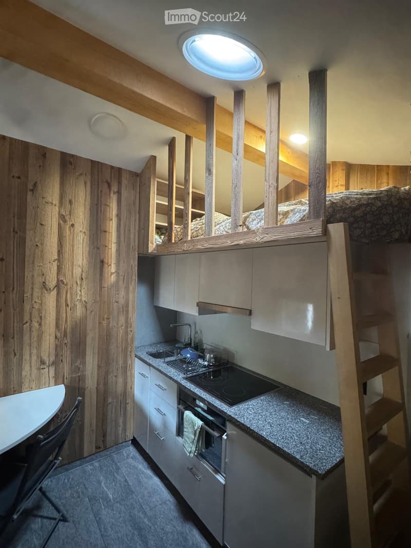 Modern kitchen with granite countertops, stainless steel appliances, and wooden beams. Features a sink, stove, oven, and a small dining area with a chair and table.