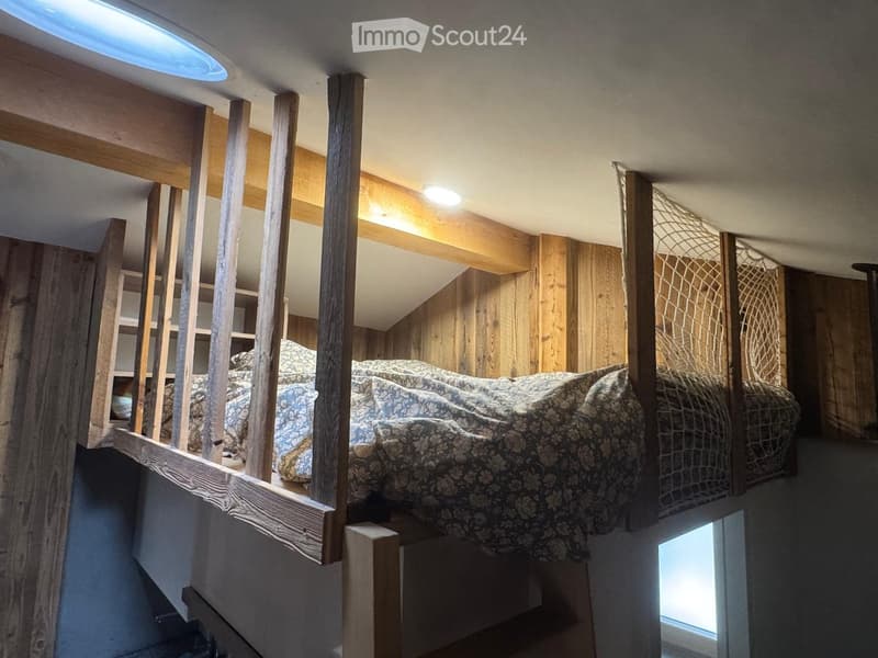 Loft-style bedroom with wooden beams, exposed wooden walls, and a raised bed platform. The bed is covered with patterned bedding and is positioned on a wooden platform with wooden railings.