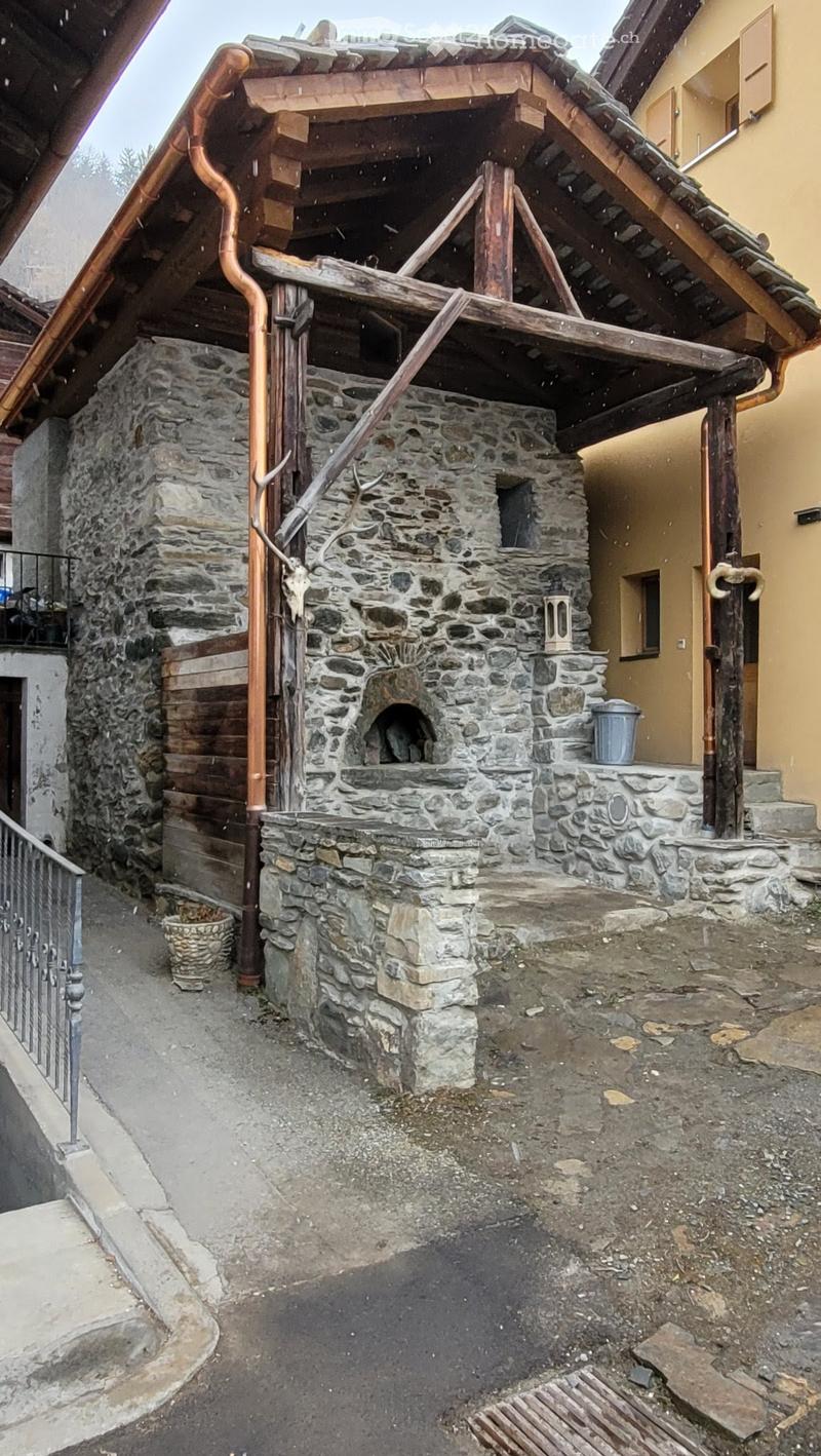 Stone outdoor structure with a raised platform, wooden beams, and a stone oven. The area includes a metal railing, a small basket, and a metal container.