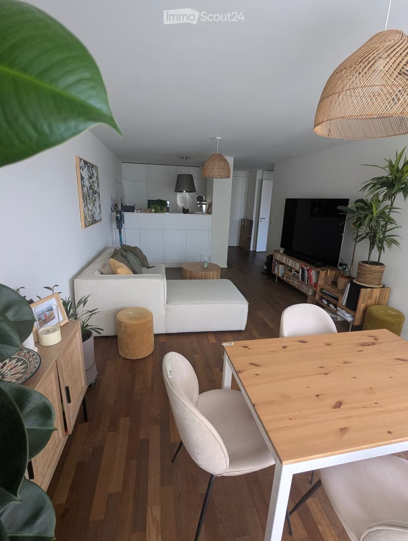 Modern living room with a white sofa, ottoman, round wooden coffee table, and a dining table with chairs. Features a kitchen area with white cabinets and pendant lights, along with a TV and bookshelf.