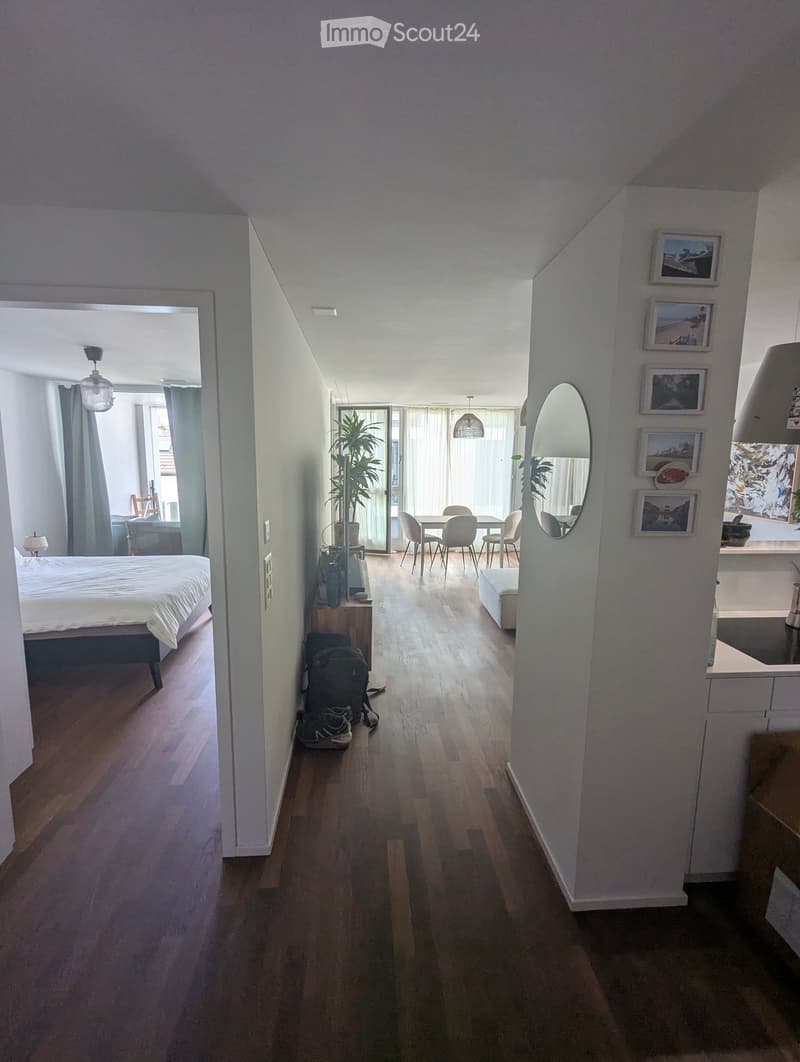 A modern hallway with wooden flooring, leading to a bedroom and a dining area. Features include a mirror, framed pictures, and a small plant in the corner.