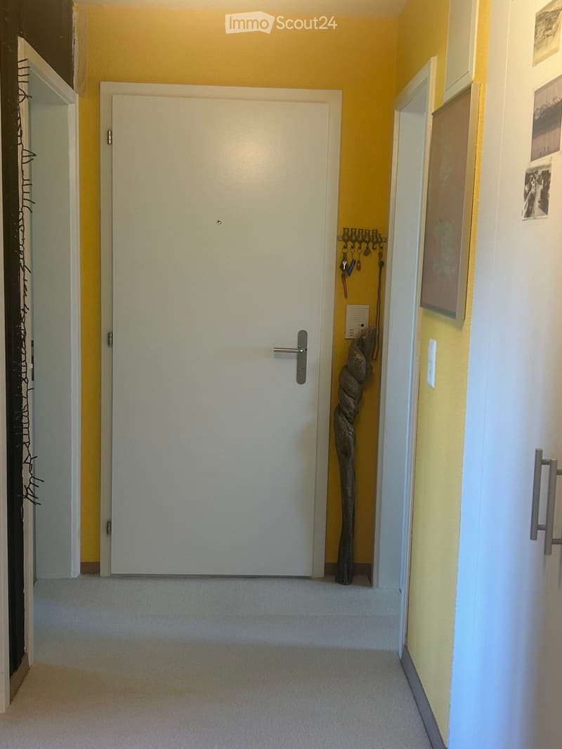 A narrow hallway with light yellow walls, carpeted floor, and white doors. A decorative wooden sculpture is placed near the wall on the right side.