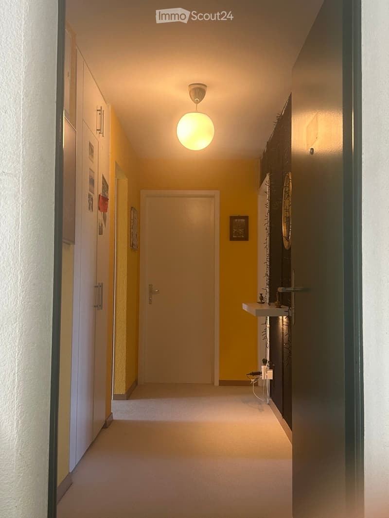 A narrow hallway with yellow walls, white carpeting, and a single round ceiling light. There are doors on both sides, a small shelf with decorative items, and framed pictures on the walls.